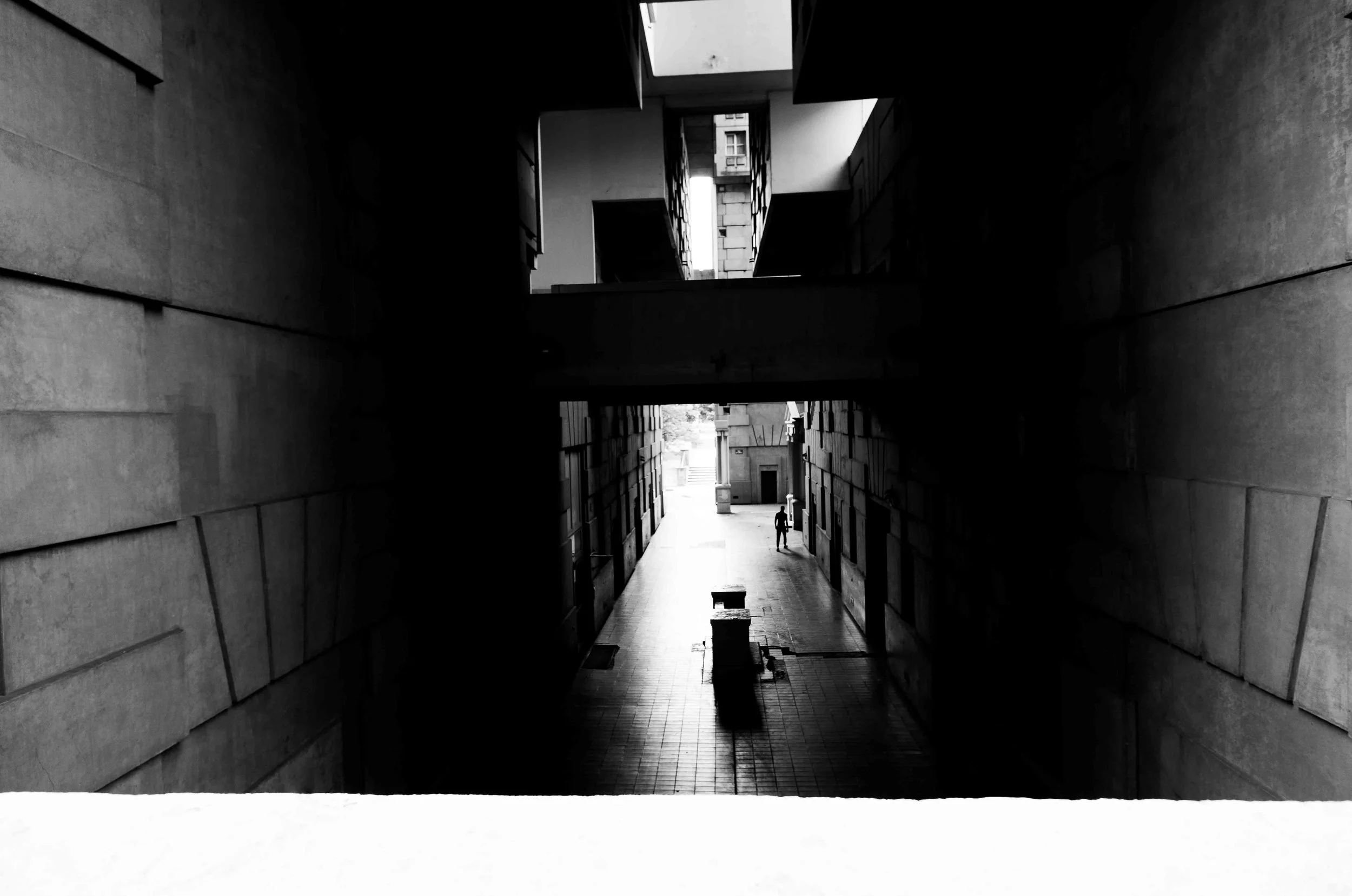 Black and white photo of a narrow city alleyway viewed from a shadowed archway, with a person walking in the distance.