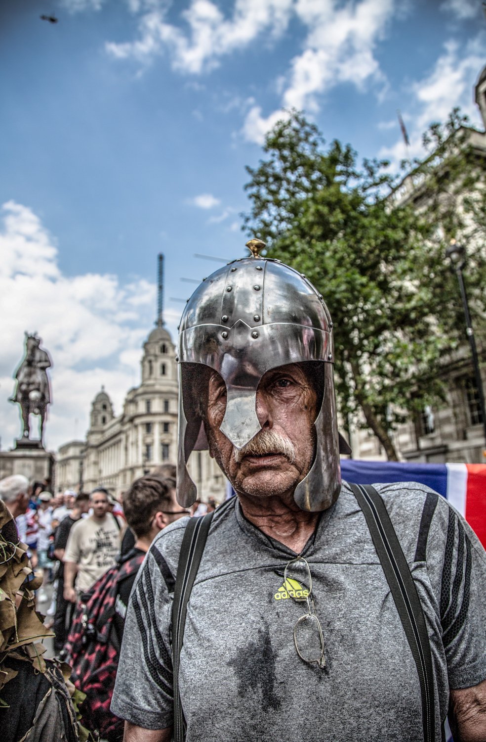 A man with a mustache wearing a helmet resembling a Roman centurion's helmet at a crowded outdoor event in a city square with historical buildings and statues in the background.