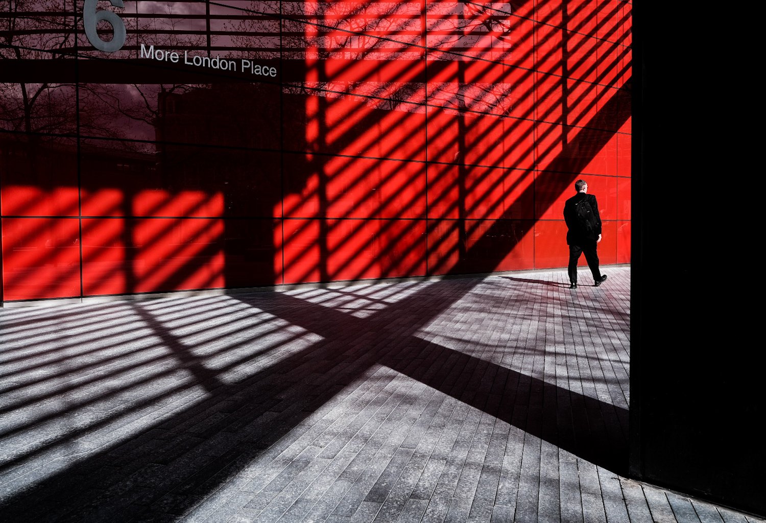 A person walking past a red building with shadows of a geometric metal structure cast on the wall and ground.