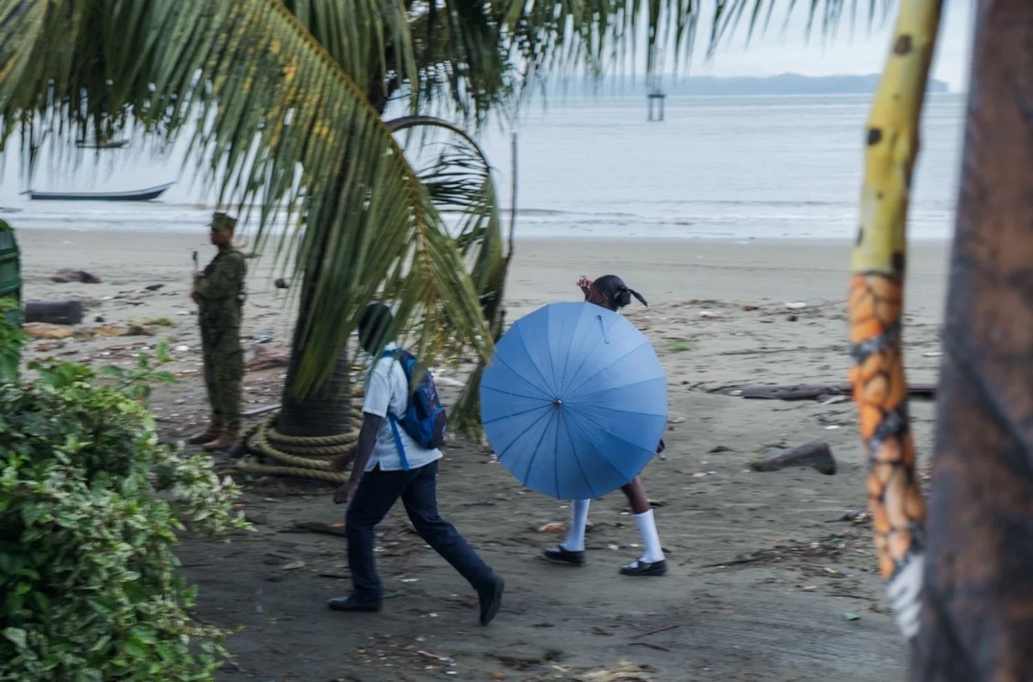 People walking on a sandy beach near the ocean, with one person holding a blue umbrella, and a man in uniform standing near a palm tree.