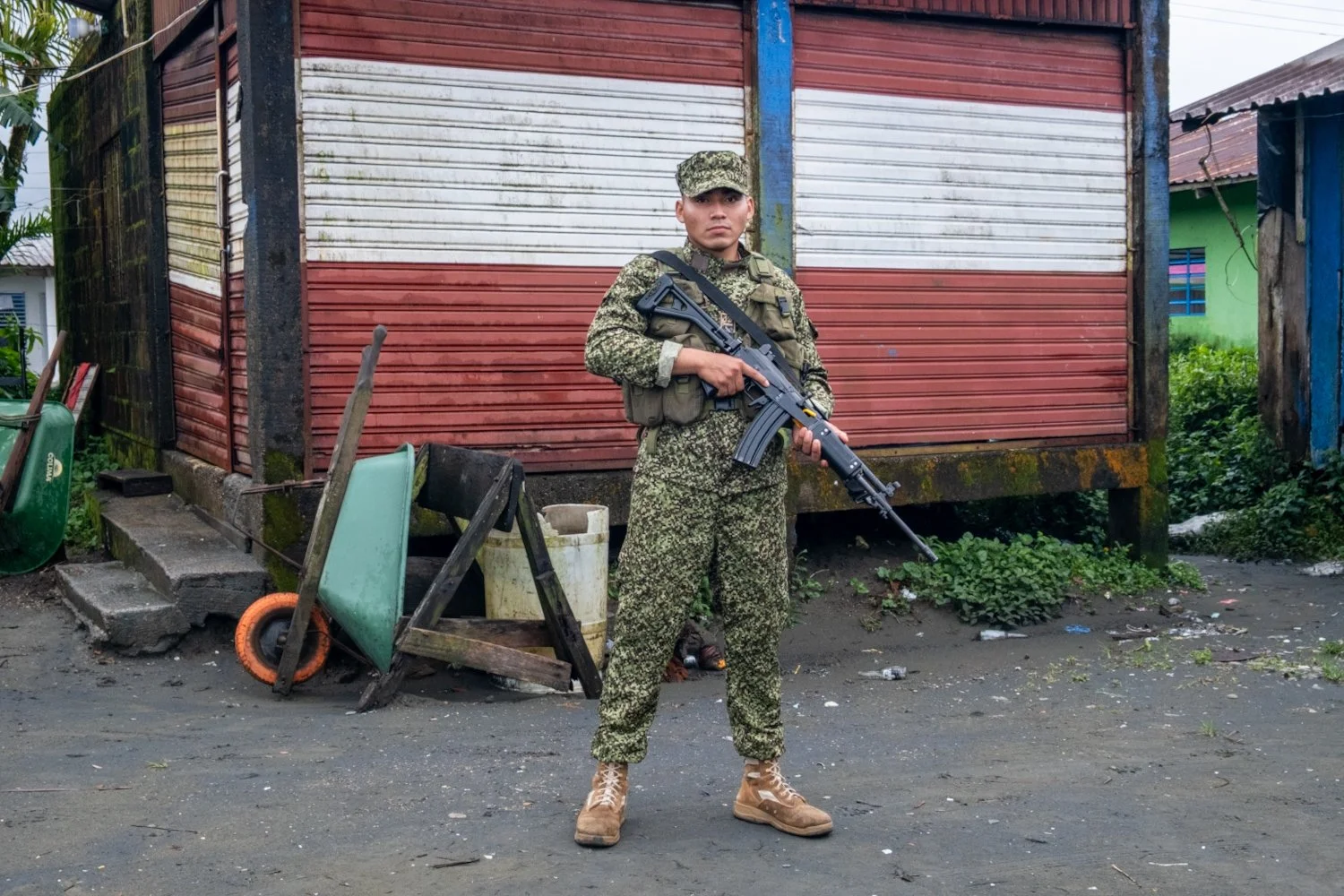 A soldier standing in front of a weathered red and white metal building, holding a rifle, wearing camouflage military uniform and tan boots, with debris and foliage around.