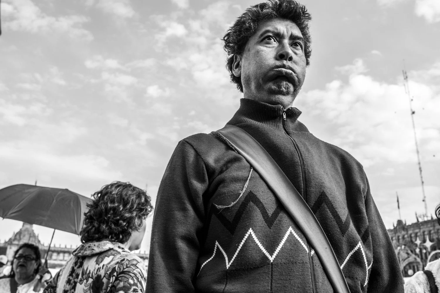 A black and white photograph of a man with curly hair and a serious expression, standing outdoors during the day. He is wearing a zip-up sweater with a patterned design and has a strap across his body. There are multiple people in the background, one