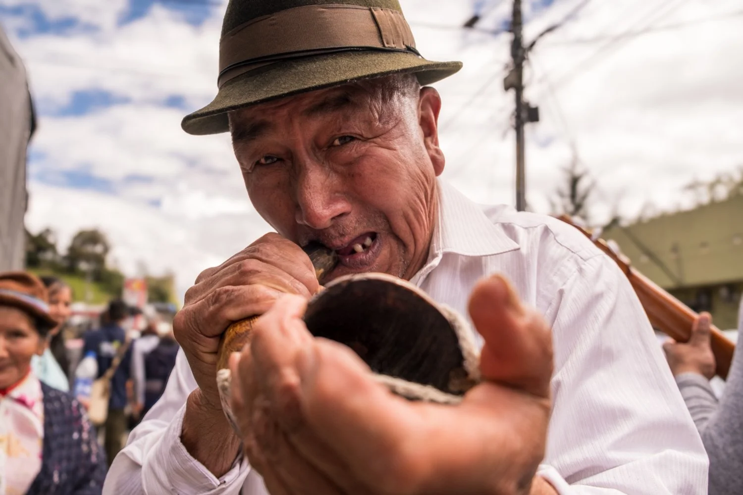 An elderly man wearing a beige fedora hat and white shirt, playing a wooden flute outdoors during daytime, with a crowd and cloudy sky in the background.