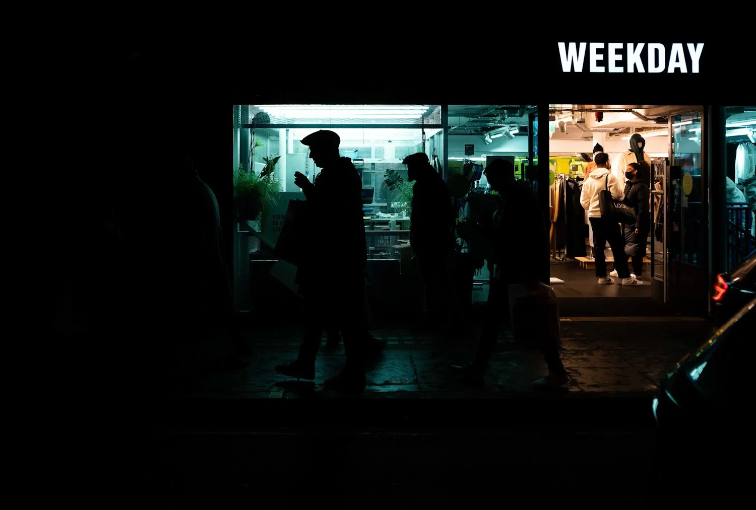 Silhouettes of people walking past a brightly lit store at night, with the word 'WEEKDAY' displayed above the entrance.