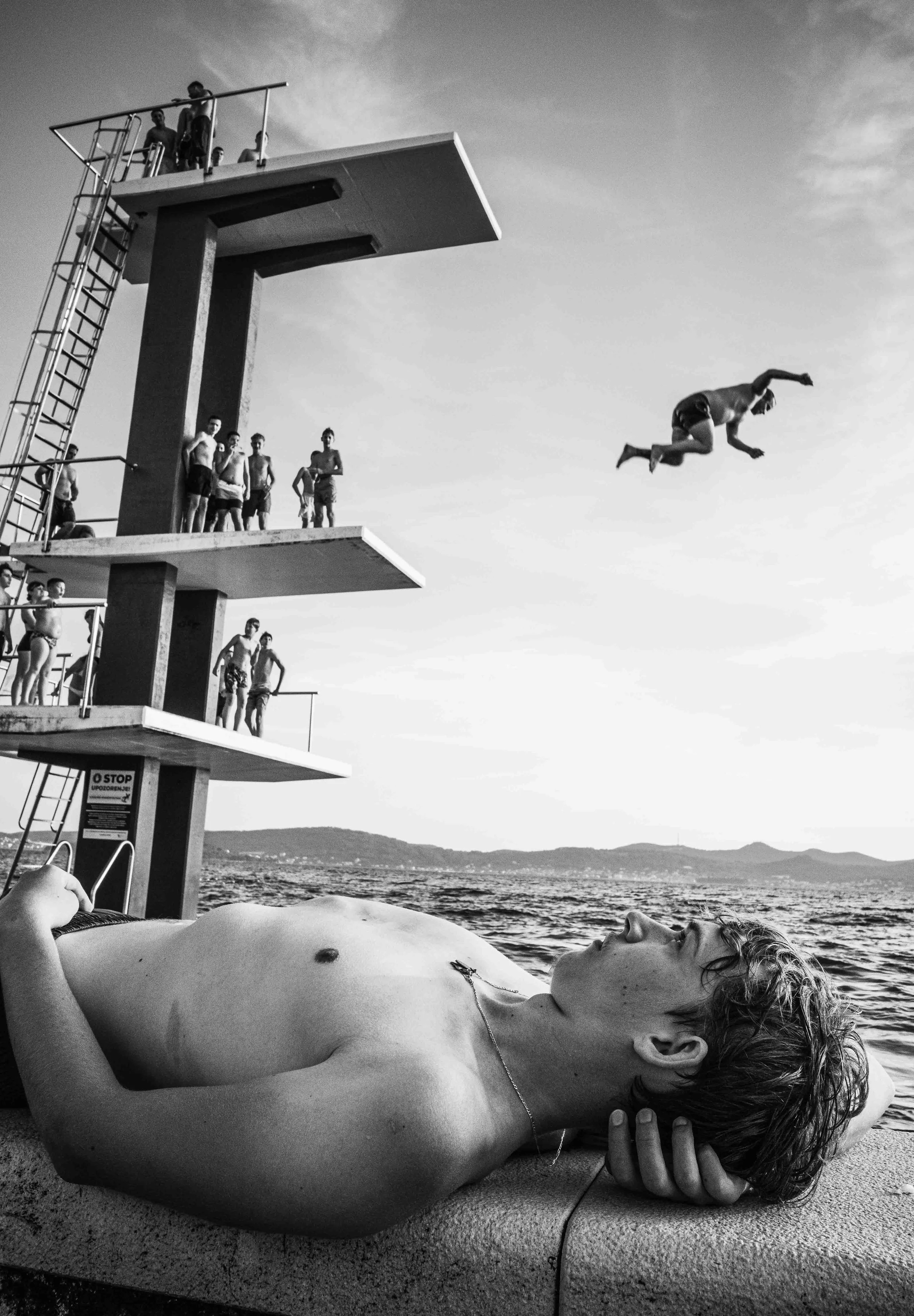A young man with wavy hair and no shirt is lying on his back on a towel by the water, relaxing with eyes closed. In the background, a diving platform with several people standing on it overlooks the water, and one person is mid-air, diving into the w