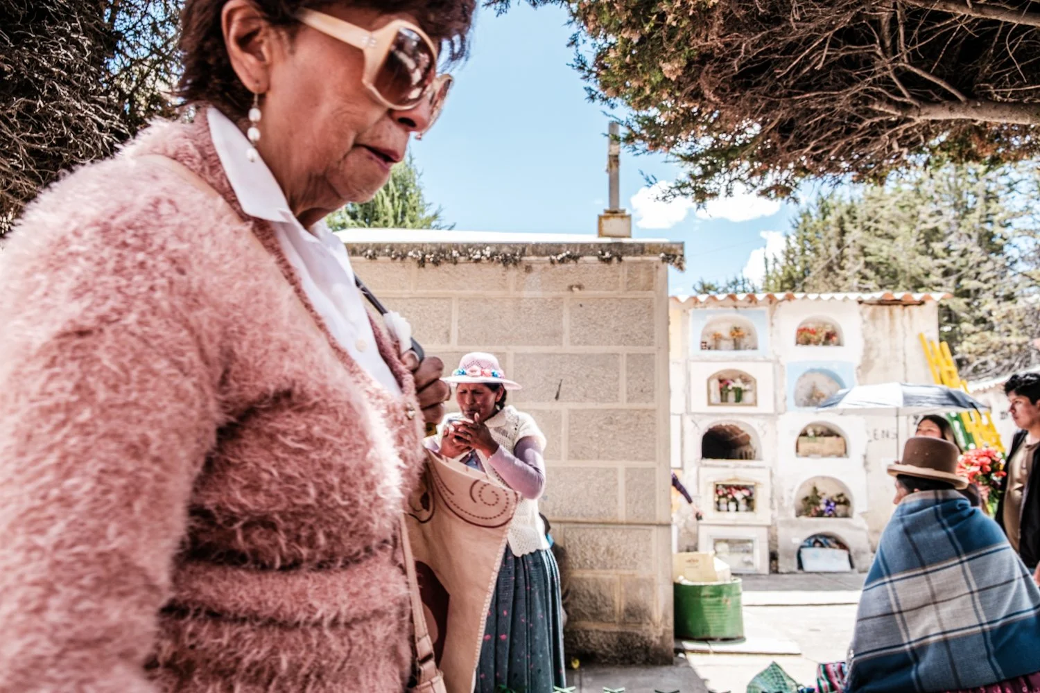 Elderly woman in pink coat and sunglasses at a cemetery with other people, some wearing traditional clothing, and a wall of niches with flowers, on a sunny day.