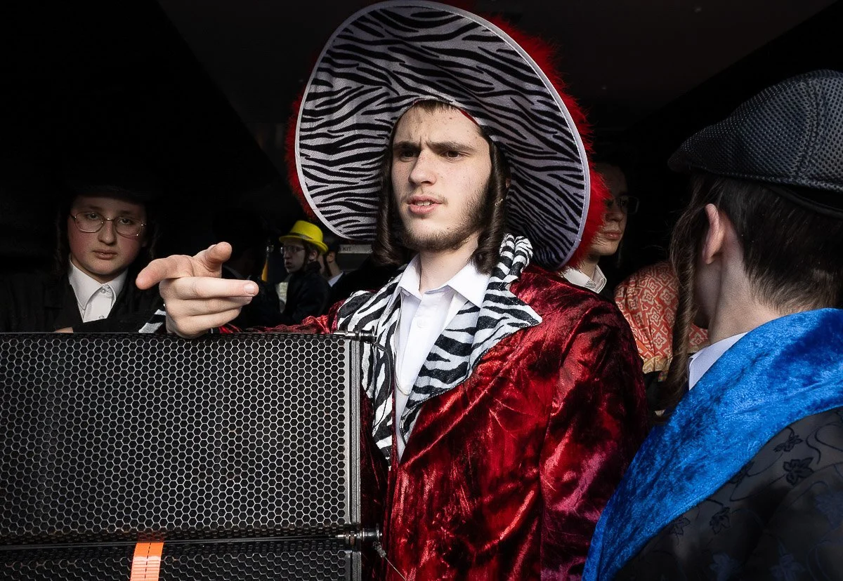 A person dressed in a red velvet coat and a zebra-print hat with a large brim, standing among a group of people wearing various costumes, including some with hats and glasses, in an indoor setting.