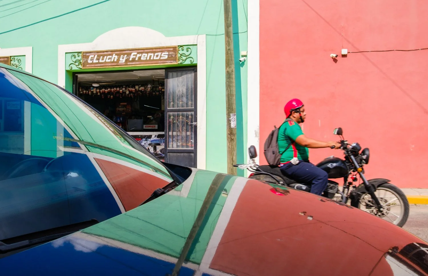 A man riding a black motorcycle past a brightly painted building with a green wall on the left and a pink wall on the right, with a storefront in the background.