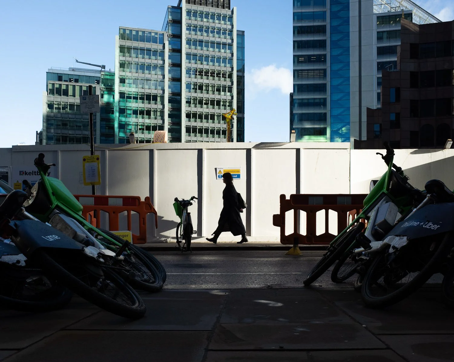 A woman walking past a construction site with a white barrier, high-rise glass buildings in the background, and several parked bikes and scooters in the foreground.