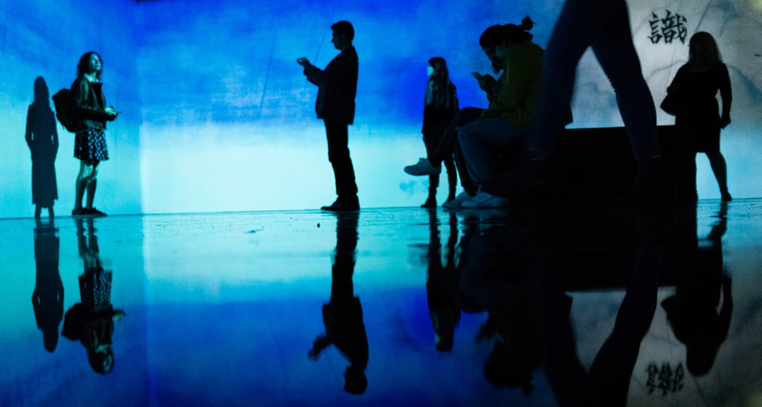 Silhouettes of people in an art exhibit or waiting area, some standing and others sitting, with a large blue-lit digital screen in the background, reflected on the shiny floor.