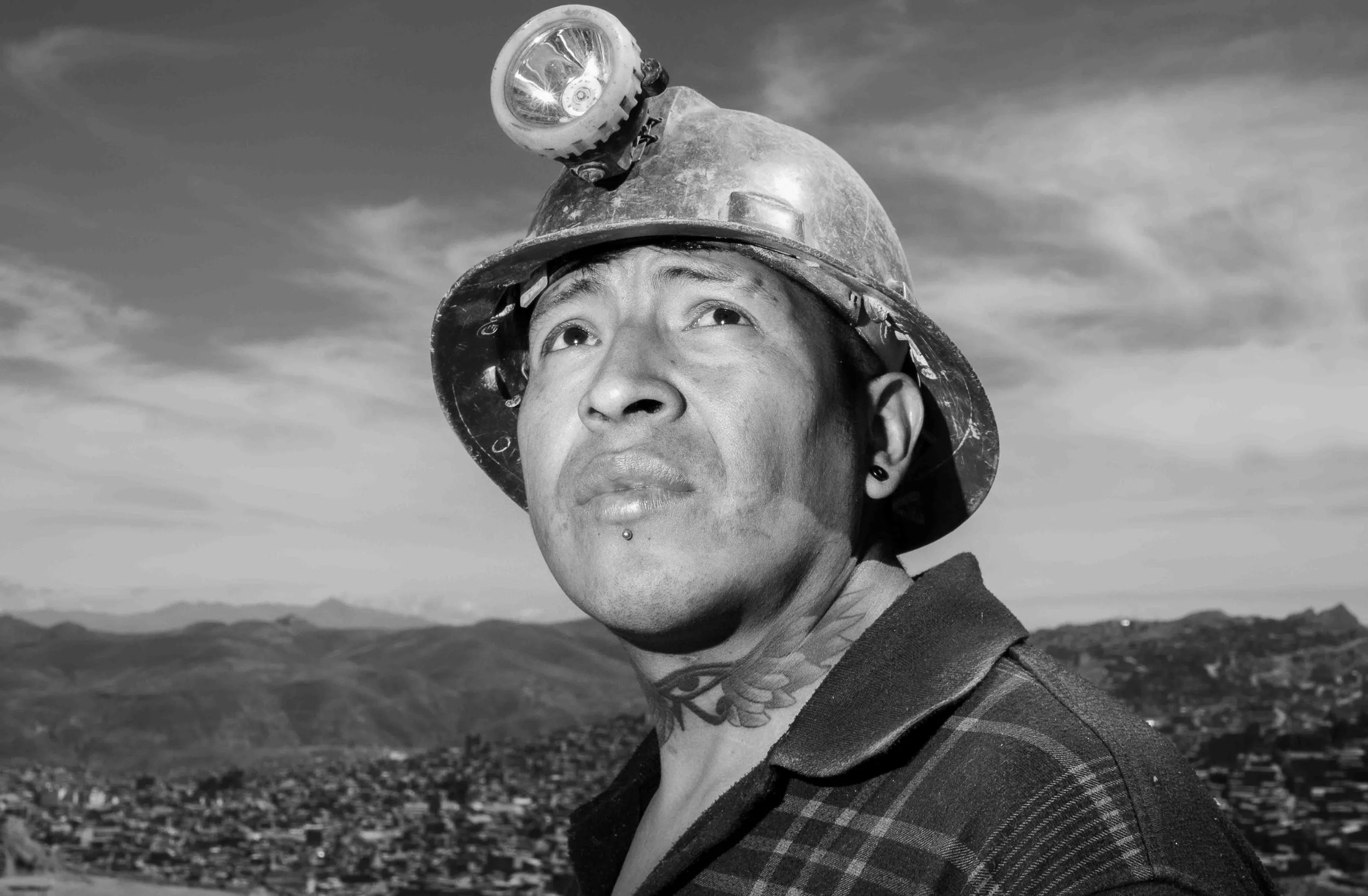 A person wearing a hard hat with a headlamp, looking upward, with a landscape of mountains and a town in the background.