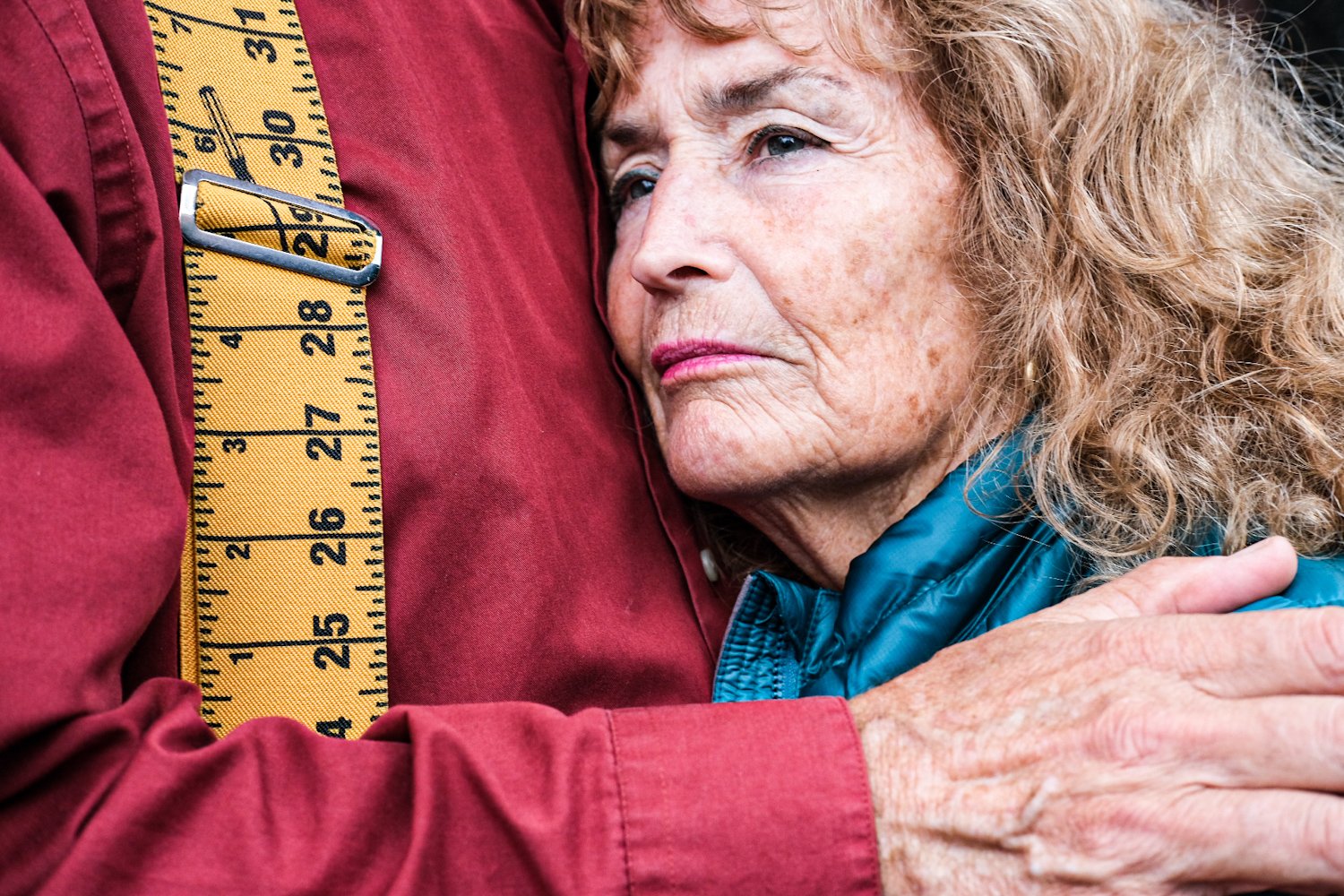 An elderly woman with curly hair hugging a person wearing a red jacket, with a yellow measuring tape over the person's shoulder.