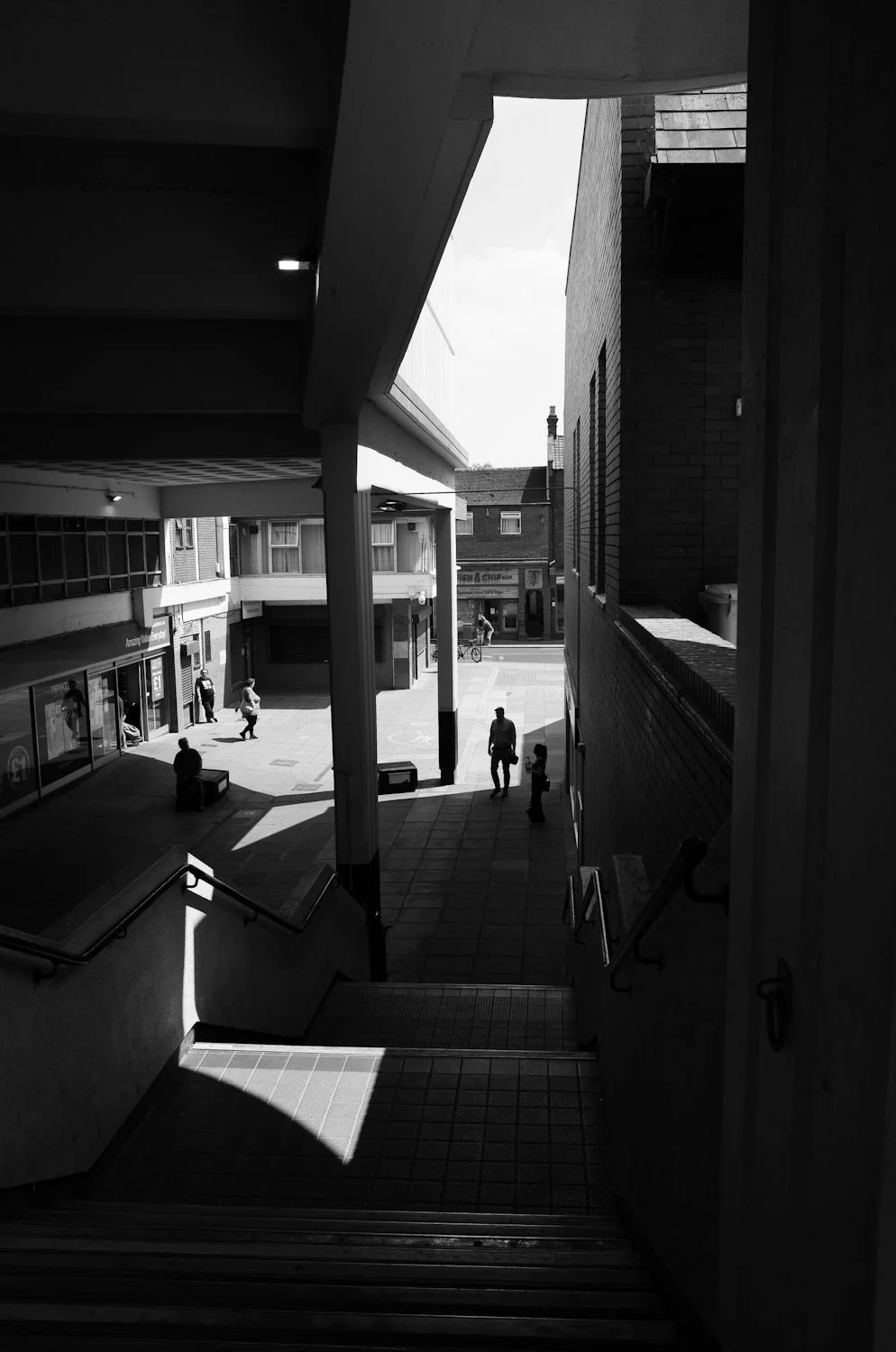 Black and white photo of an urban shopping area with stairs leading down to a street, showing pedestrians and storefronts.
