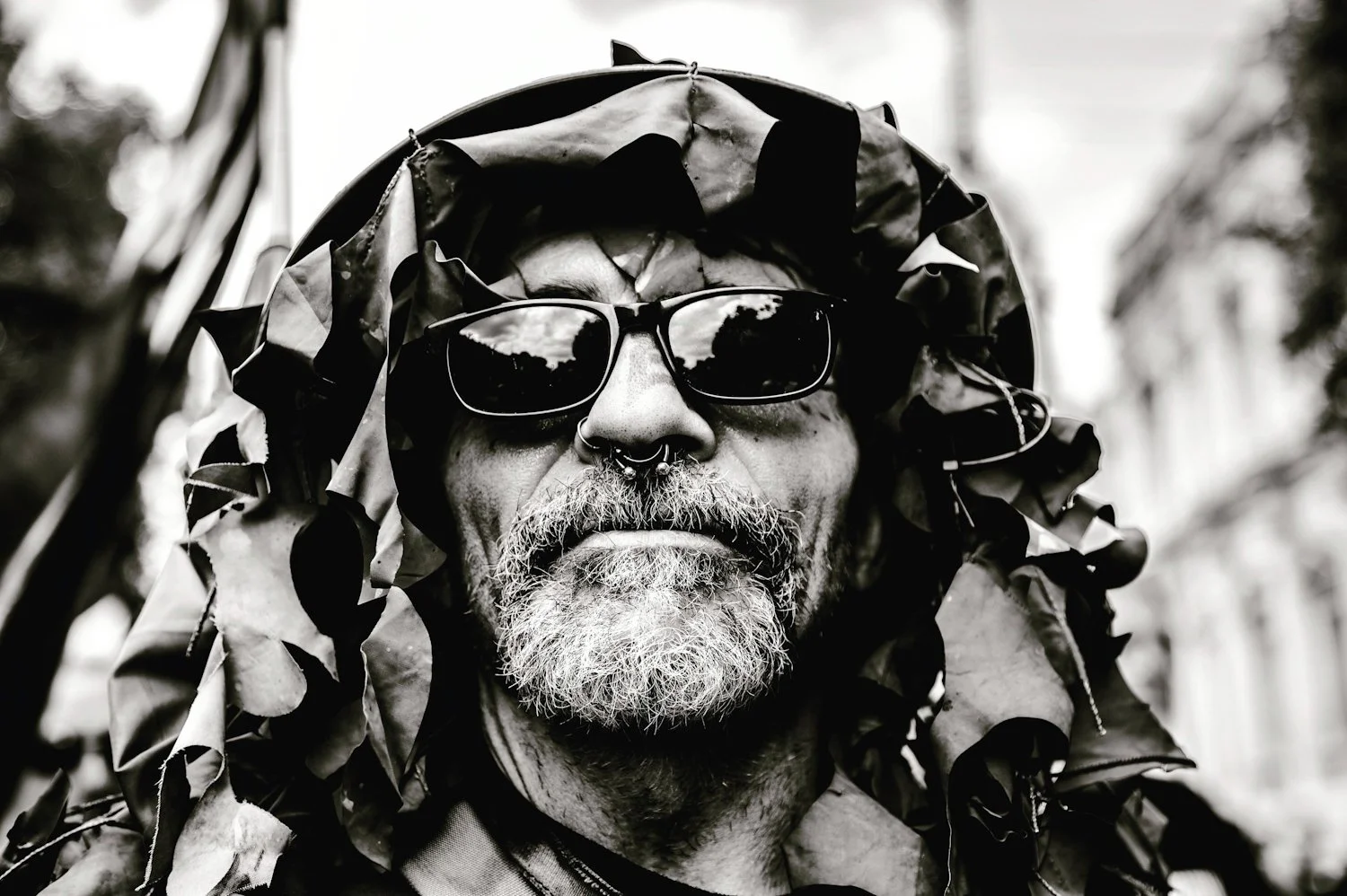 A black and white close-up photograph of a man with a rugged appearance, sporting sunglasses, a septum piercing, and a beard, wearing a camouflage-like hood made of leaves and branches.
