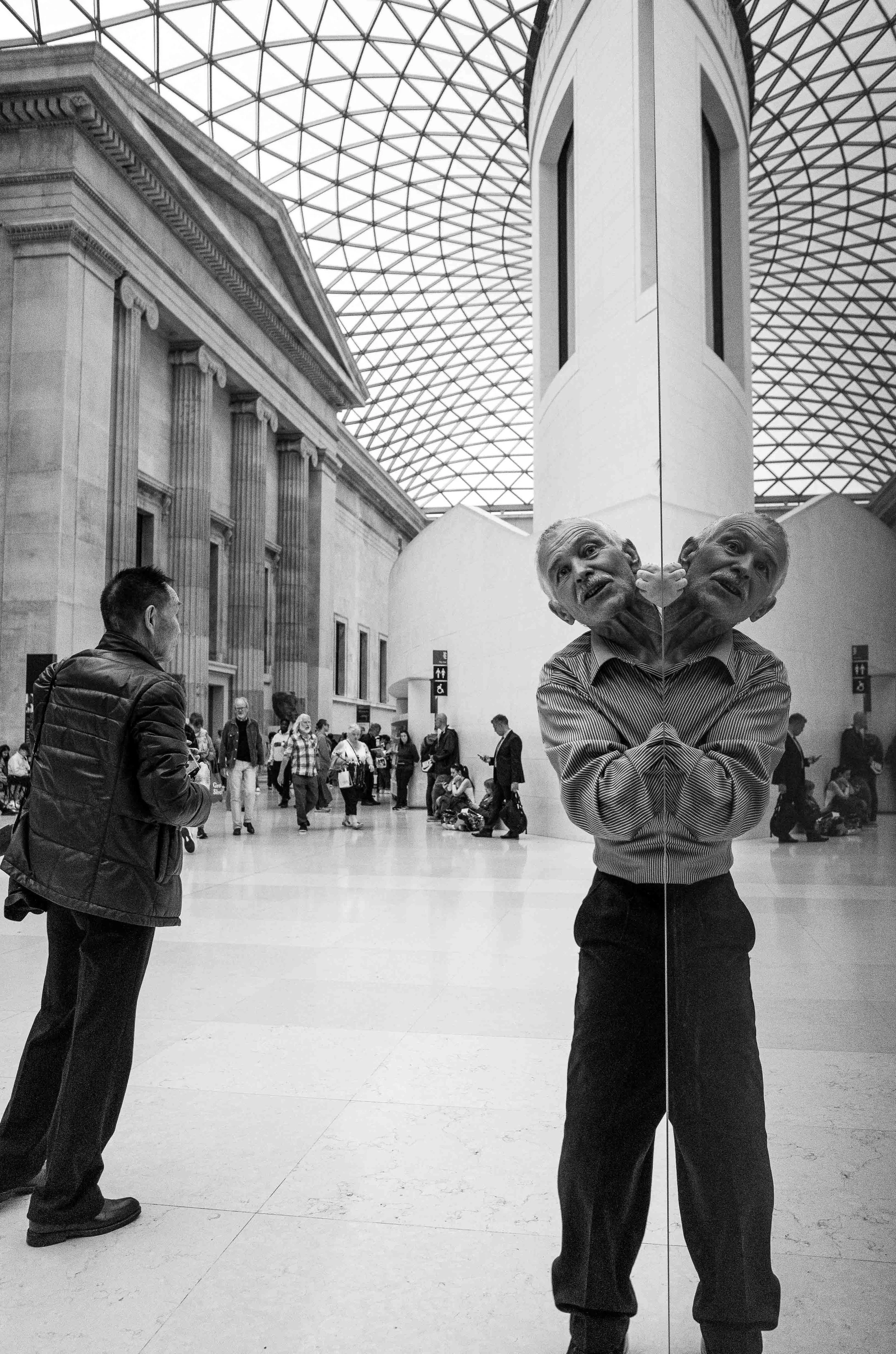 A man taking a photo in a large indoor space with a high arched glass ceiling. The photo captures the reflection of his face on a mirrored wall that has a matching reflection of his face. Other people are visible in the background, some walking and o