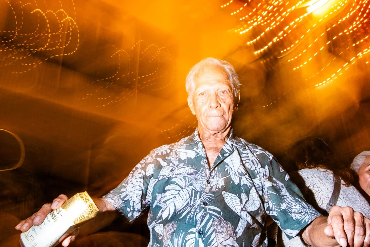 An elderly man in a Hawaiian shirt holding a drink, with a colorful, festive background of blurred lights.