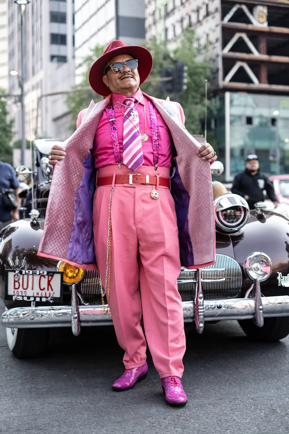 Person dressed in bright pink outfit, including suit, tie, shoes, and accessories, standing in front of a vintage black car with a license plate marked "BUICK," on a city street with buildings and spectators in the background.