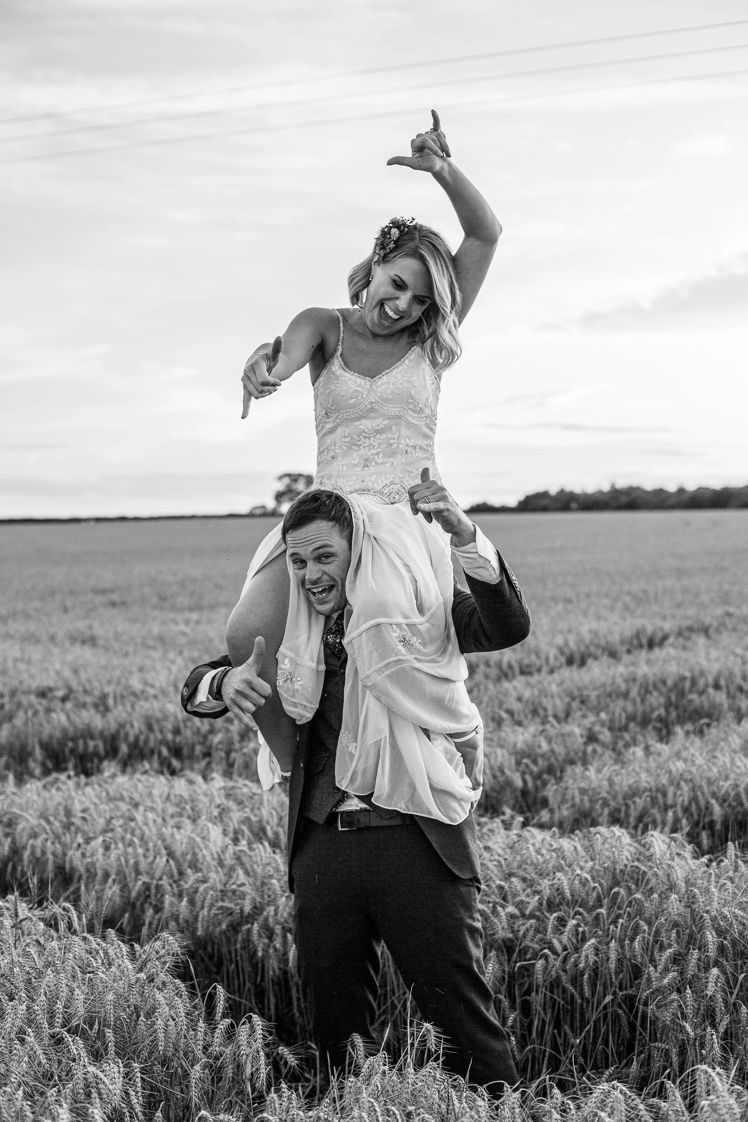 A woman in a wedding dress sitting on a man's shoulders while standing in a wheat field, smiling with her arm raised in celebration.
