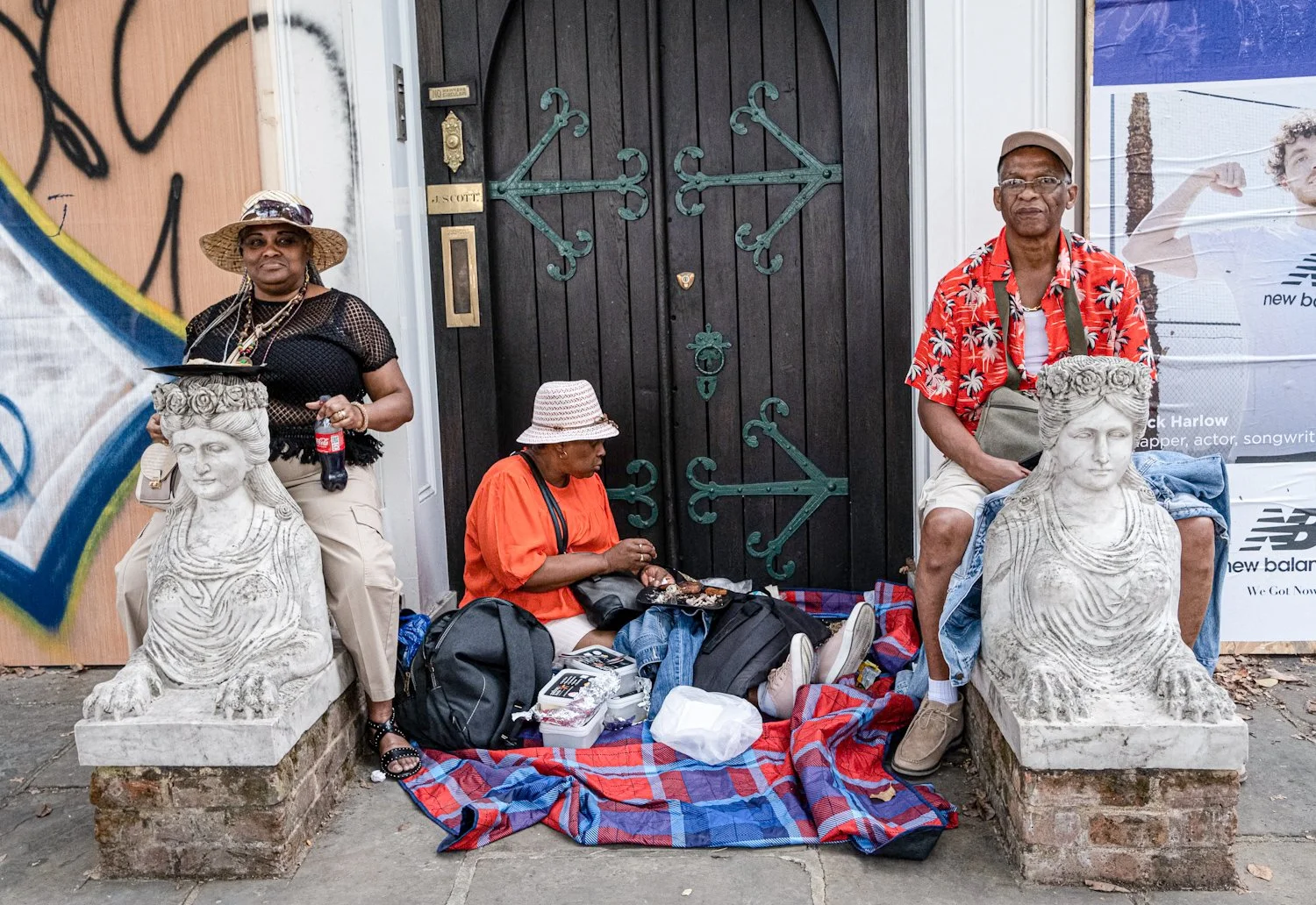 Three people sitting and standing by a doorway with black wooden doors, two stone lion sculptures, and graffiti on a wall. One woman sits on the ground with food and bags, while a man and woman stand nearby, dressed casually.