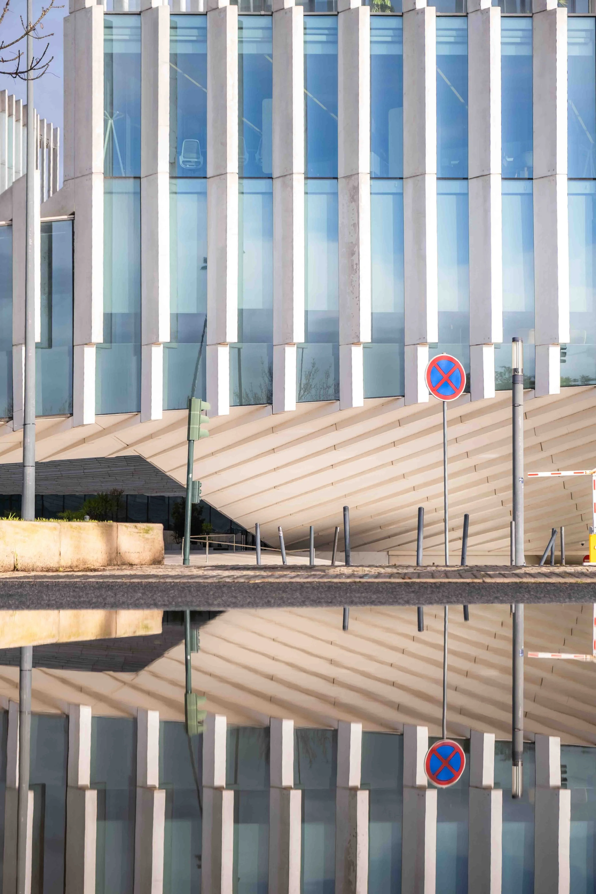 A modern building with a glass facade and vertical concrete stripes, with a 'no parking' sign and street elements in the foreground.
