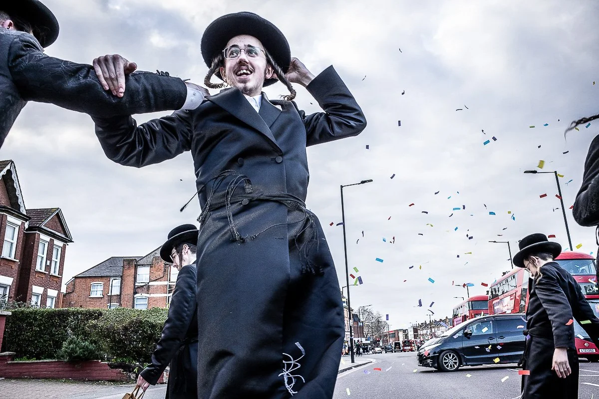 A person dressed in black with glasses and dreadlocks celebrates on a street with confetti, others around are also in black, some wearing hats, and there are cars, houses, and red buses in the background.