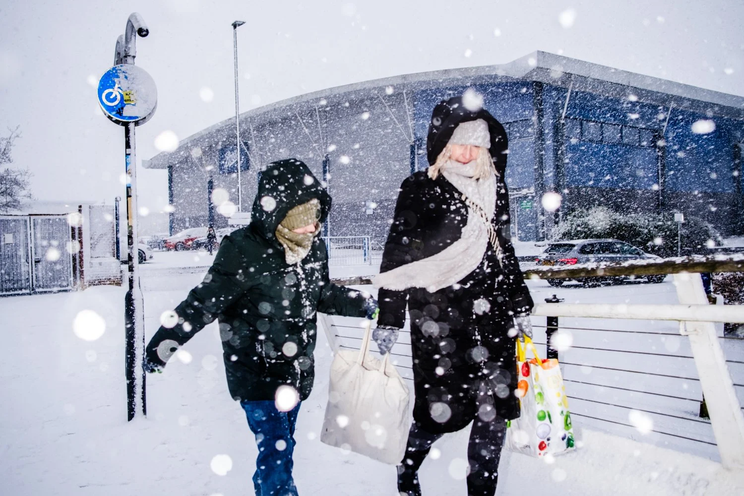 Two people walking in heavy snowfall, bundled up in winter clothing, carrying shopping bags outside in front of a modern building.