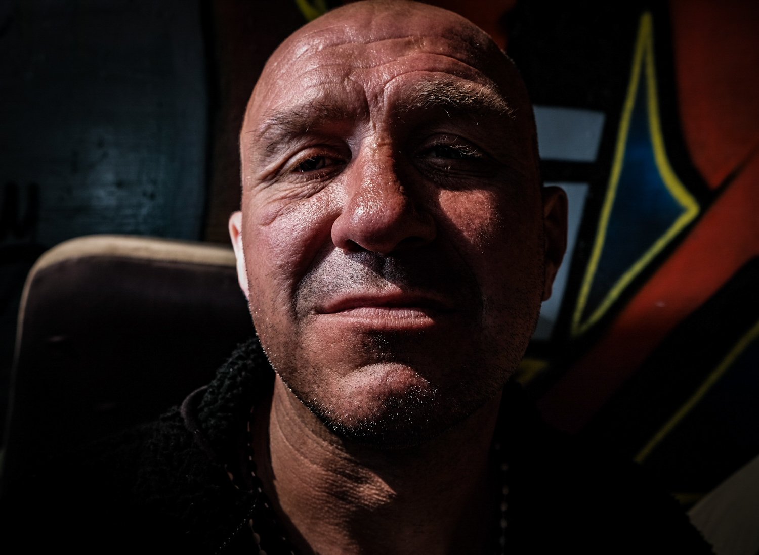 Close-up of a middle-aged man with short hair, slightly squinting, in dim lighting with a dark background that includes a partial yellow and black star symbol.