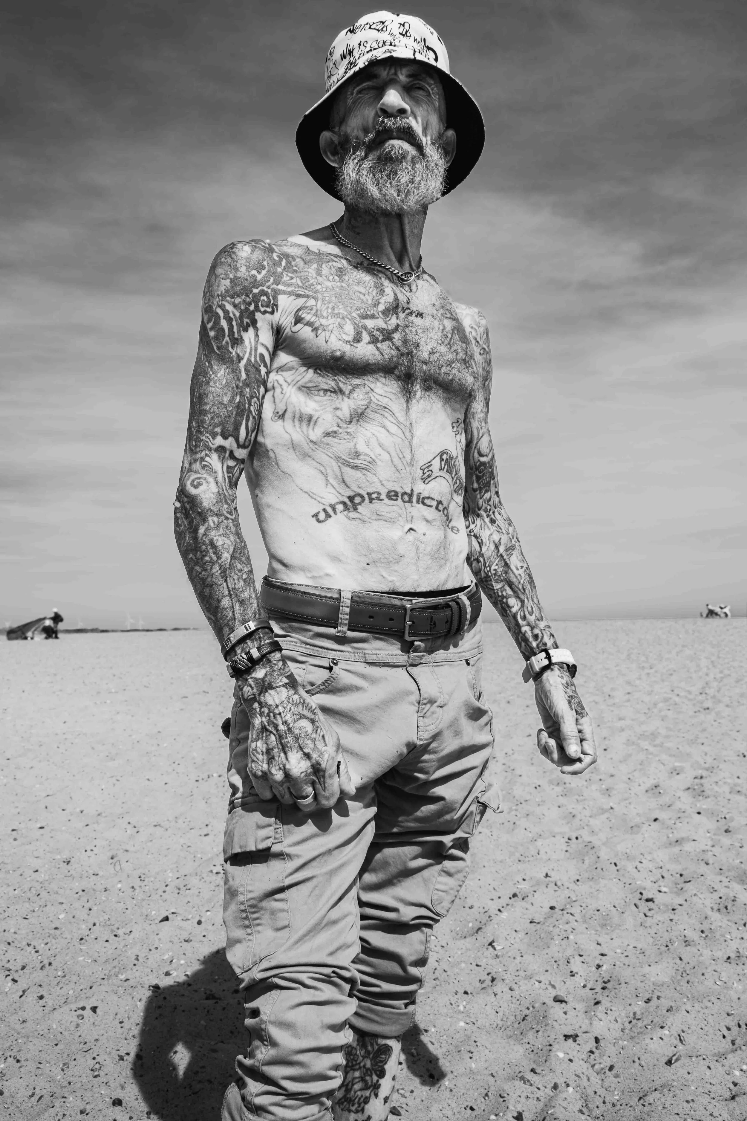A shirtless man with numerous tattoos, a beard, and a hat, standing on a beach in black and white. He has a serious expression and is looking off into the distance. There are horses in the background.