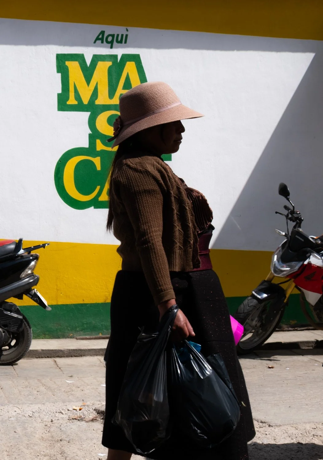 Silhouette of a woman wearing a sun hat and holding a black plastic bag, standing in front of a colorful mural with green, yellow, and white sections and the text "Aquí M A C".