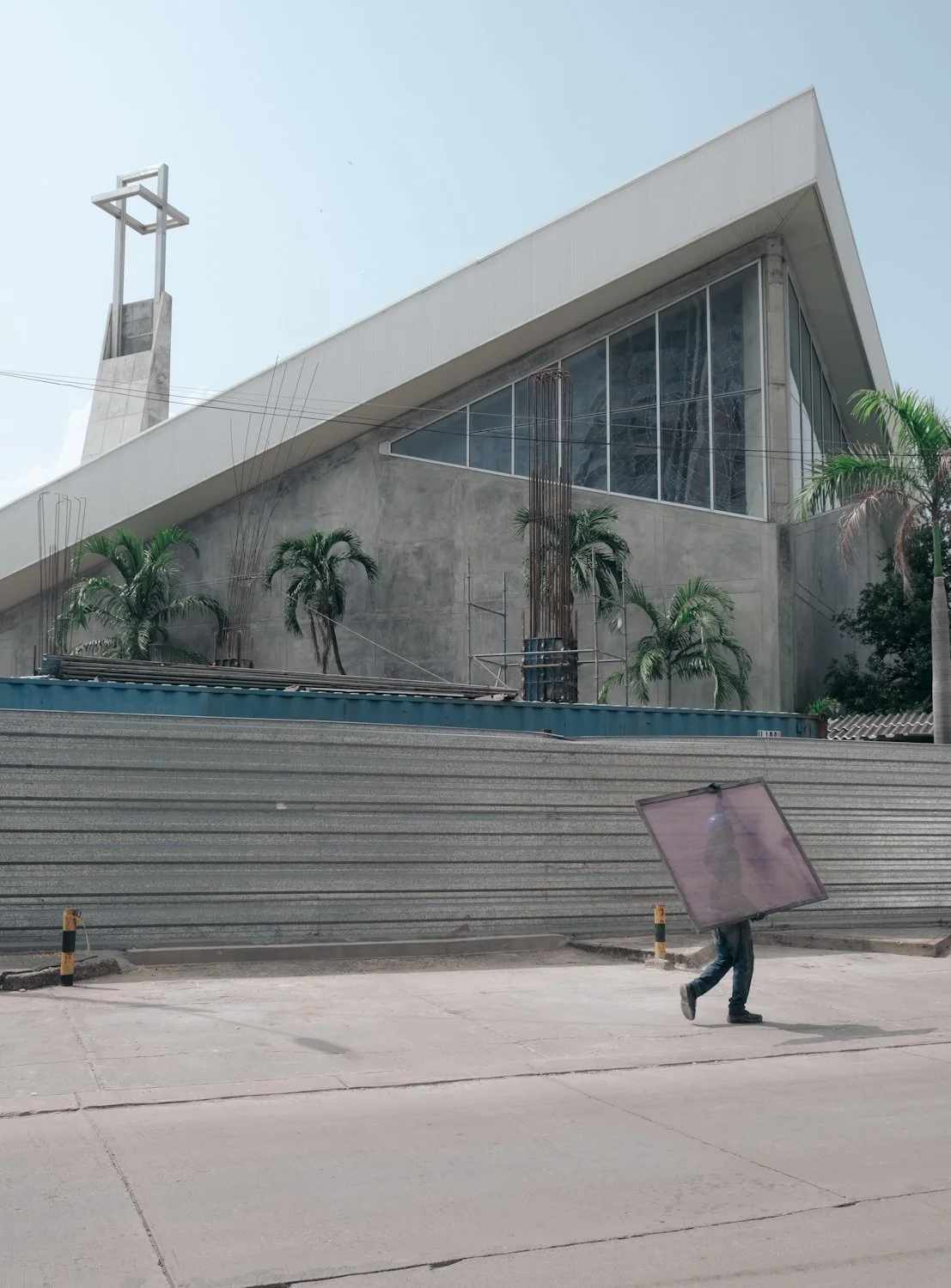 Person walking on sidewalk carrying a large pink reflective panel, with a modern building and palm trees in the background.