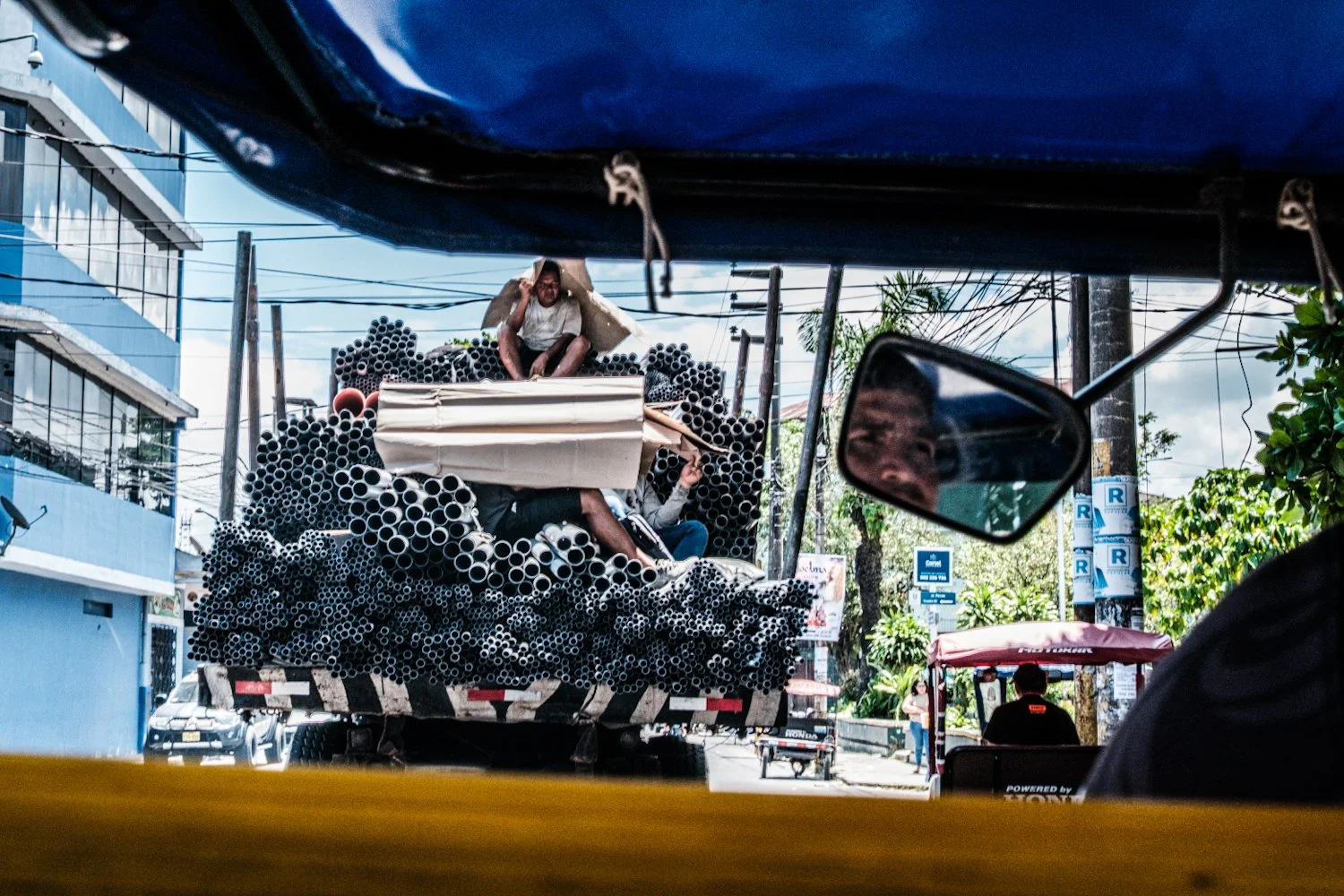 A truck loaded with black pipes, with a man sitting on top, is seen from inside a vehicle, with a side mirror reflecting part of a person's face and a woman in the background on the street.