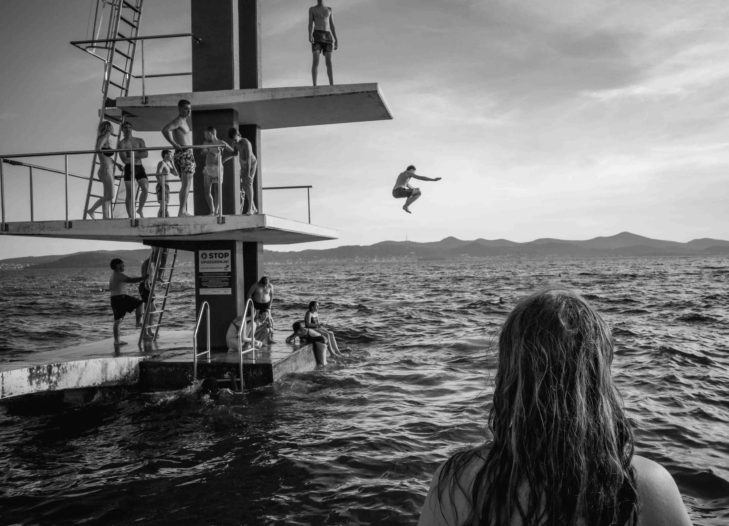A person with long hair sitting by the water, watching a diving platform where people are preparing to jump into the water, with a diver mid-air in the background.