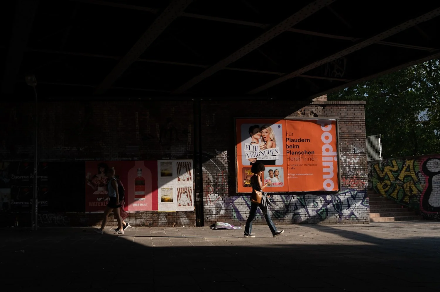 A street scene with two women walking in sunlight, graffiti-covered brick wall, and an orange billboard with German text and images of people.
