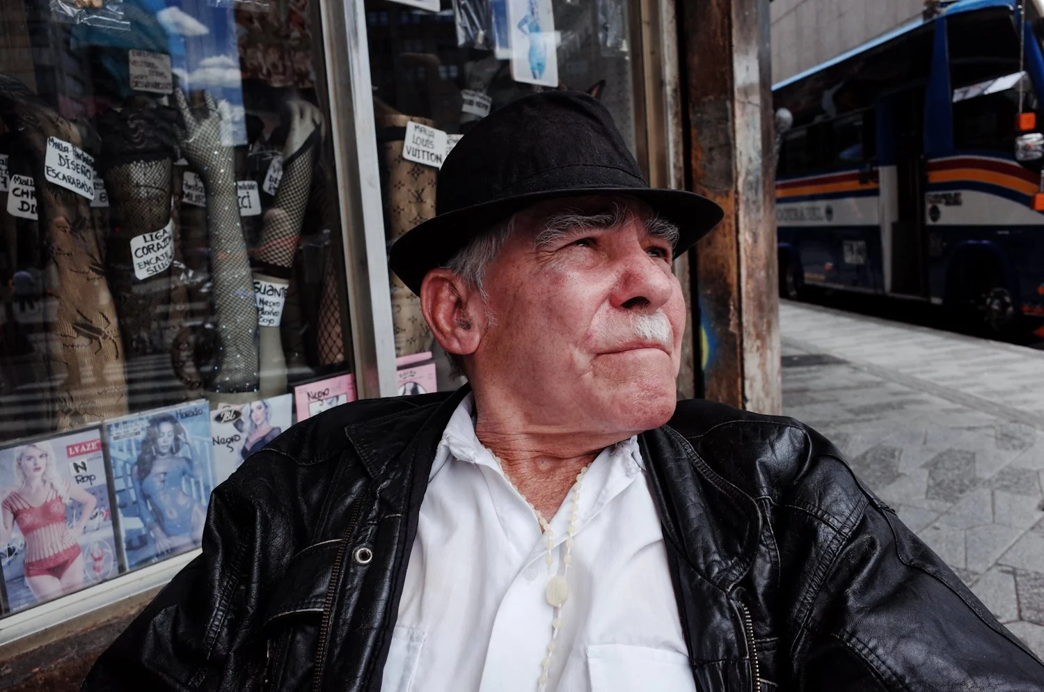 An older man with gray hair wearing a black hat, black leather jacket, and white shirt with a shell necklace, sitting on a sidewalk outside a shop window.