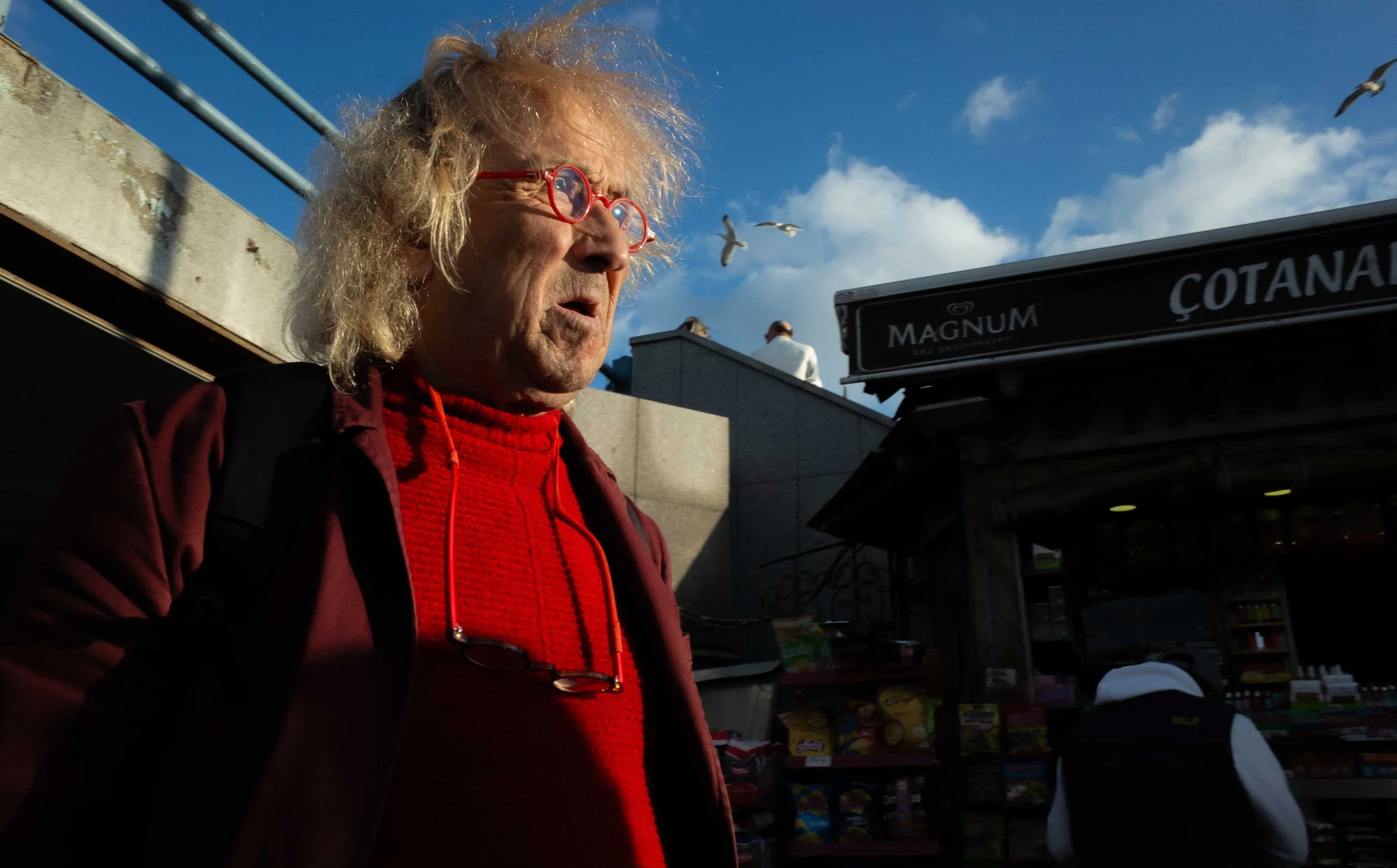 An elderly man with curly blonde hair and red glasses is standing outdoors in the late afternoon sunlight, wearing a red sweater and a maroon jacket. In the background, seagulls are flying near a clear blue sky with a few scattered clouds. There is a