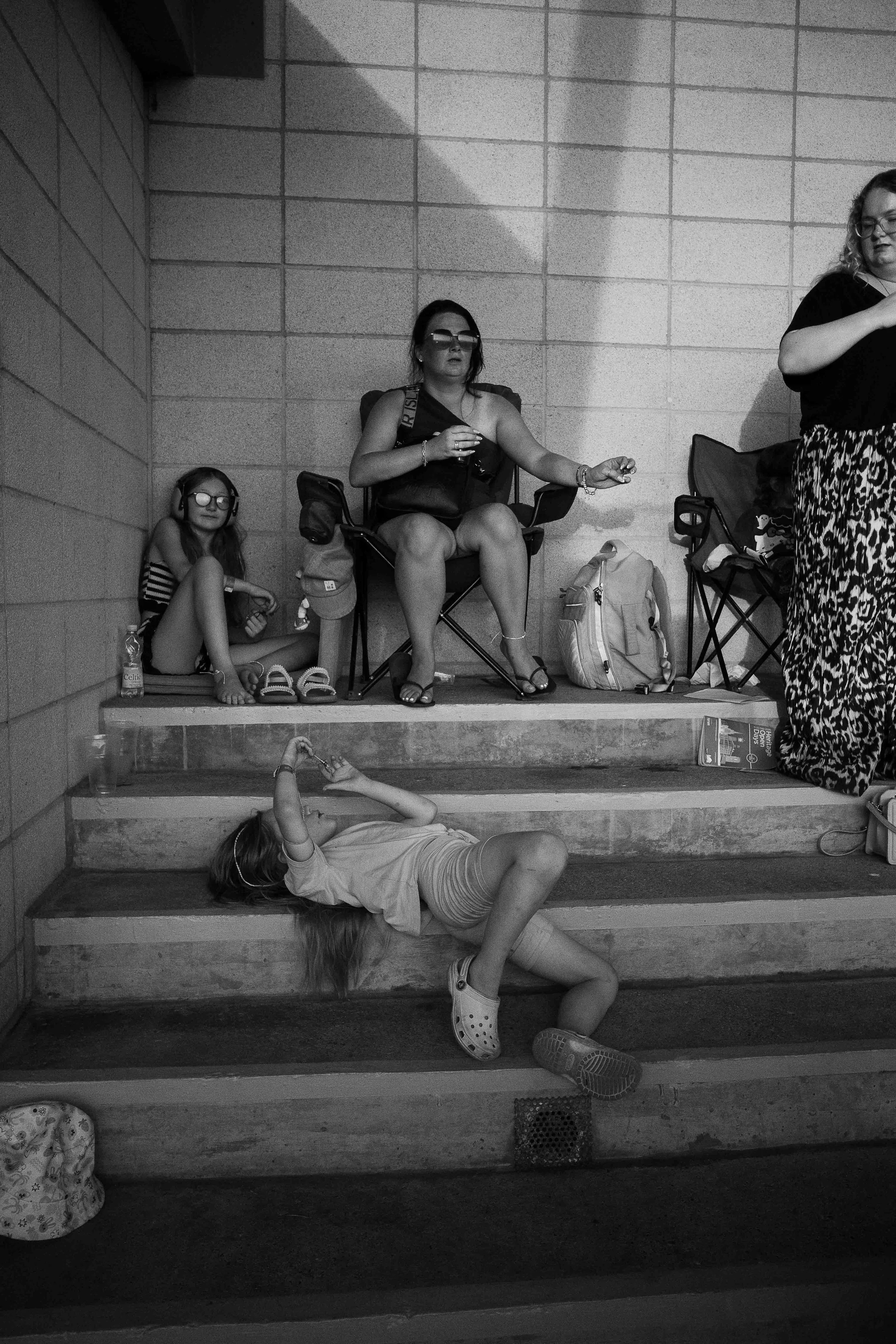 A grayscale photograph of four people in a corridor. Two are seated on chairs, and two are lying on steps below. The seated woman has glasses, dark hair, and is wearing sandals and casual clothing. The girl sitting on the ground has glasses, headphon