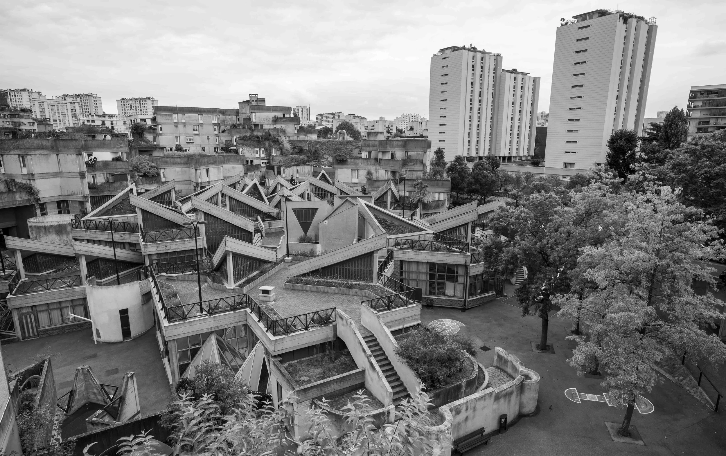 Black and white cityscape with modern multi-story apartment buildings in the background and a geometric terrace garden in the foreground, including staircases and small trees.