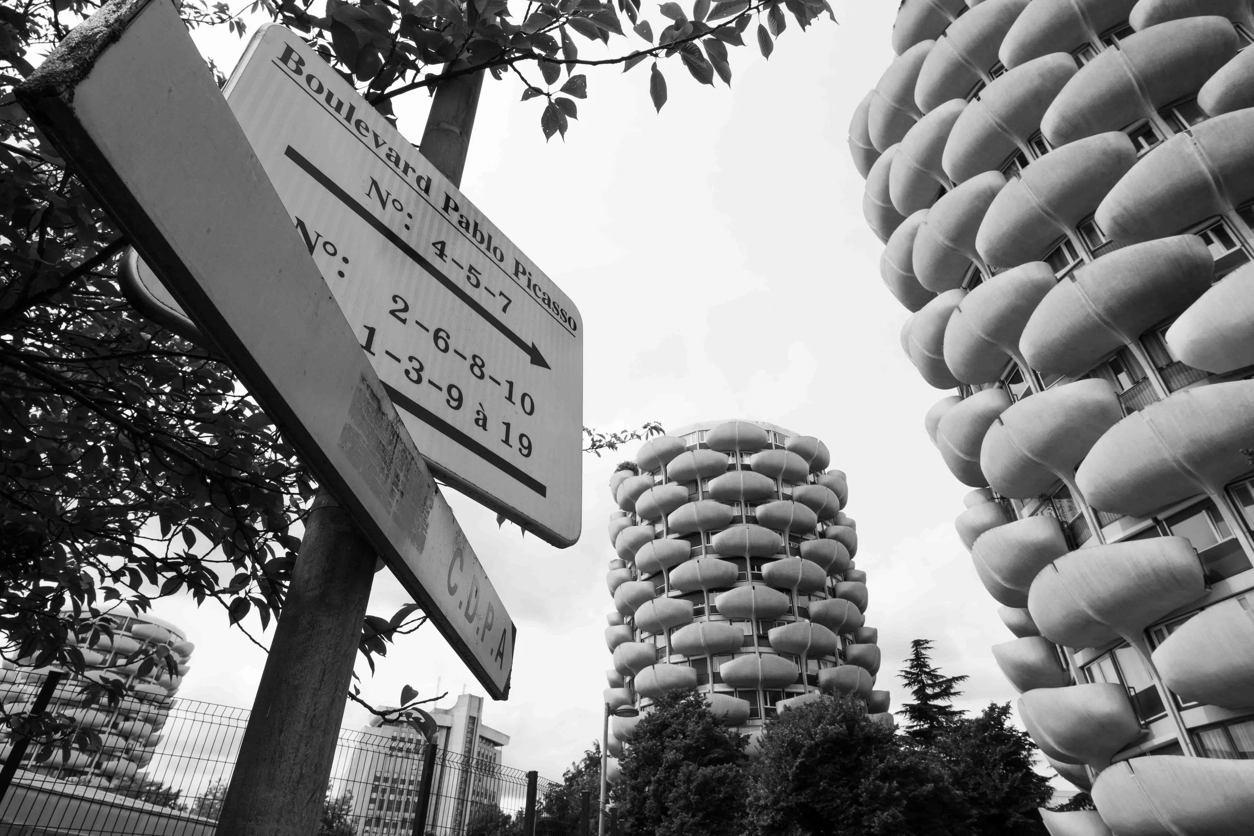 A street sign for Boulevard Arta Pablo Picasso pointing to addresses 4-5-7 with an arrow. In the background, there are tall modern residential buildings with stacked, rounded balconies and windows, along with some trees and a partly cloudy sky.