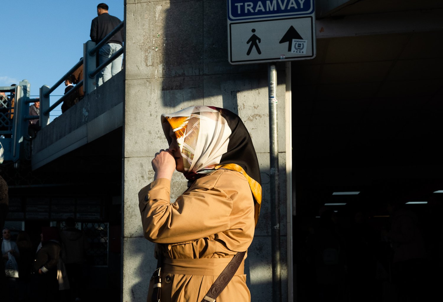 A woman standing under a bridge, wearing a beige coat and a silk headscarf, with her hand near her cheek, next to a sign for a tramway and pedestrian walkway.