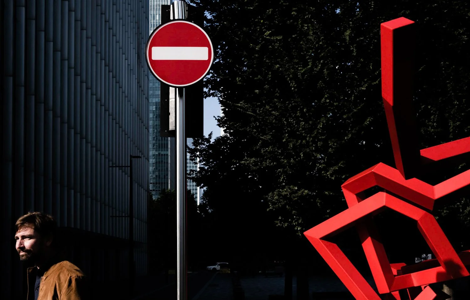 City street scene with a no entry traffic sign, a man in a brown jacket, modern buildings, and a large red geometric sculpture, with trees in the background.