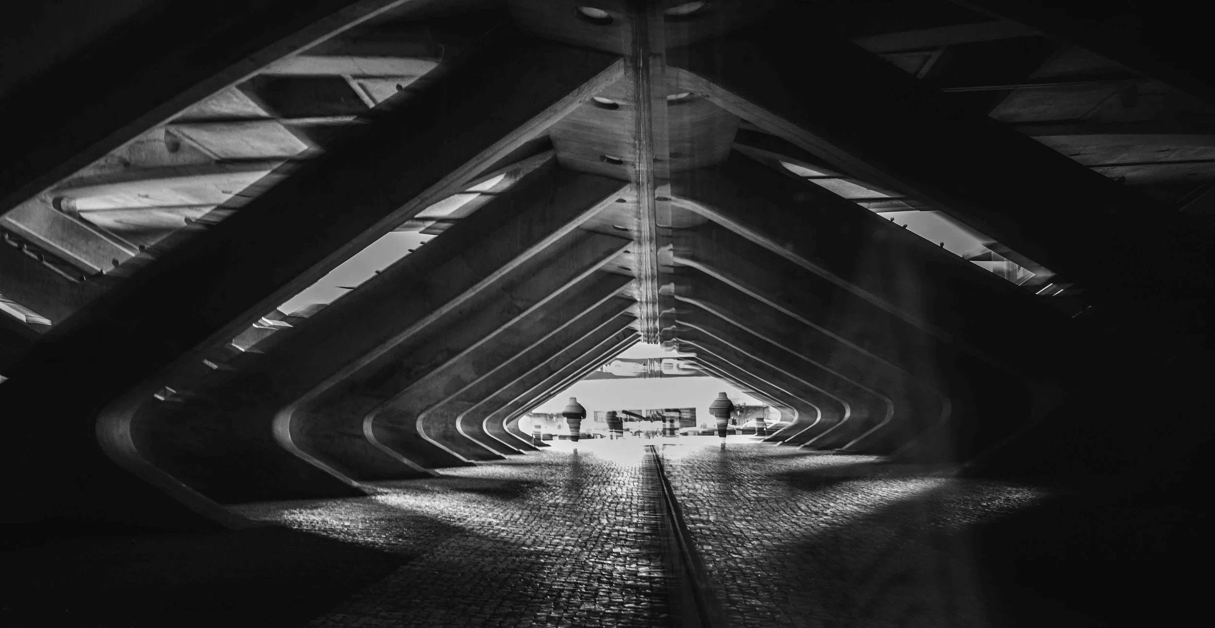 A black and white photograph of a modern architectural structure with a tunnel-like walkway. The underside features repeating geometric patterns created by angled beams. Two people are walking away from the camera towards a bright opening at the end 
