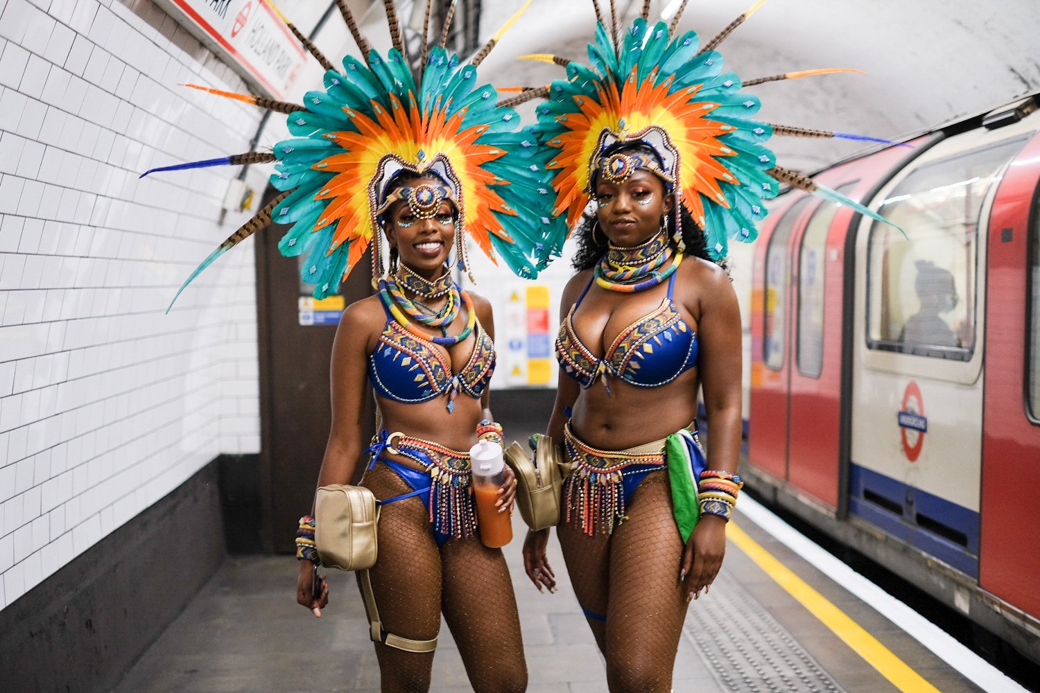 Two women in carnival costumes with large colorful feathered headdresses, beads, and fishnet stockings on a subway platform.
