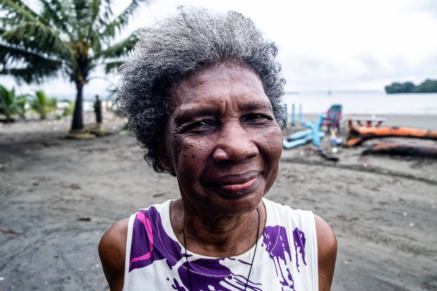 Close-up of an older woman with gray, curly hair smiling outdoors at a beach with palm trees, boats, and water in the background.