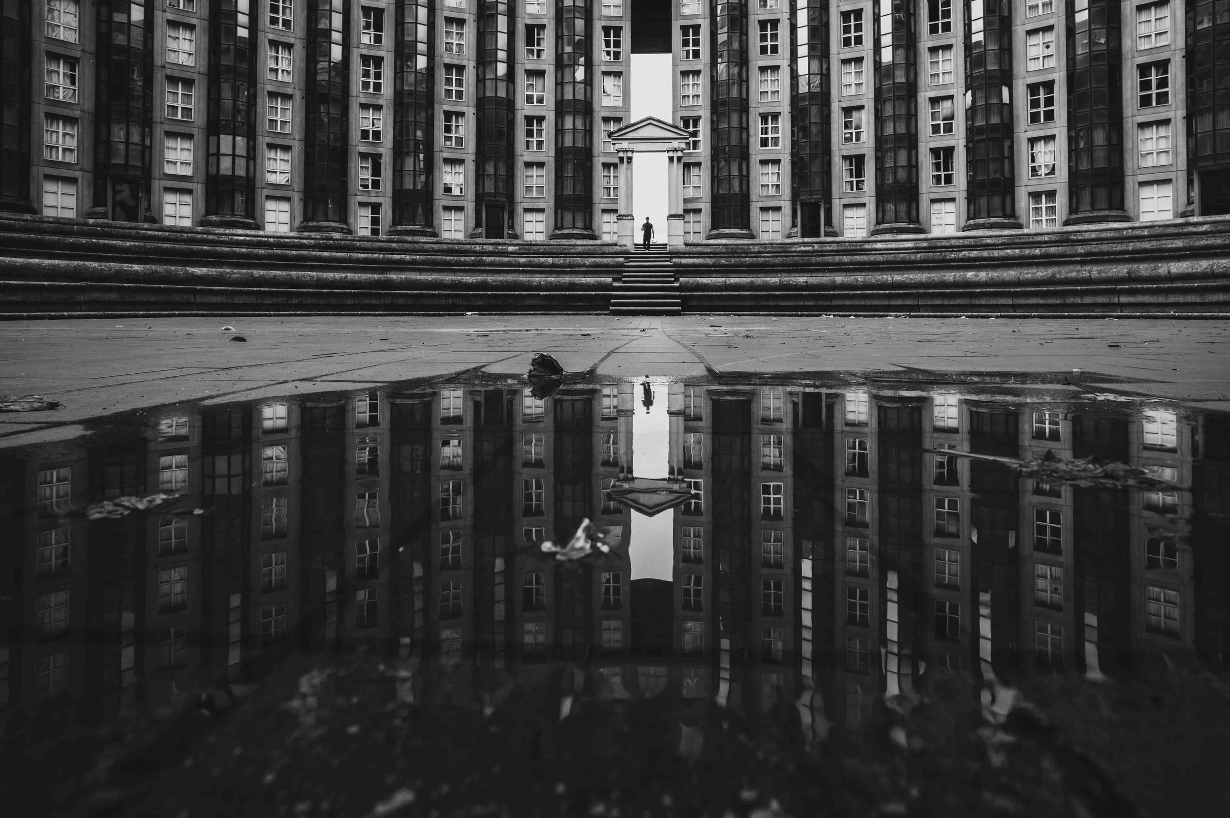 Black and white photo of a modern residential or office building with numerous windows. A set of stairs and a person are visible in the center, with a small water puddle reflecting the building and stairs in the foreground.