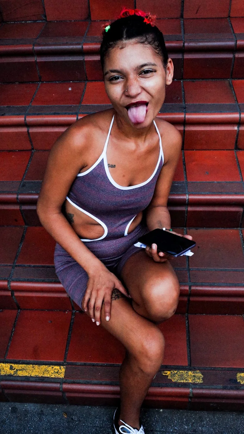 A young woman with skin tone, wearing a sleeveless gray dress with white trim, sits on red tiled steps. She has a playful expression with her tongue out, short curly hair with red and green hair clips, and a nose piercing. She is holding a phone in o