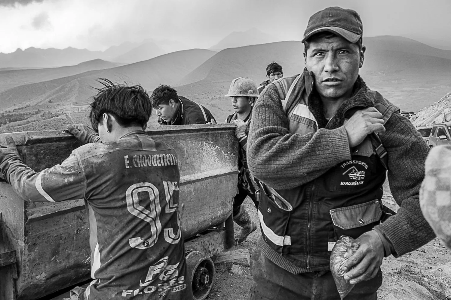 A group of people, likely workers or migrants, are seen outdoors on a dirt road with mountains in the background. One man in the foreground is looking at the camera, while others are partially visible and appear to be working or resting beside a metal cart or container.