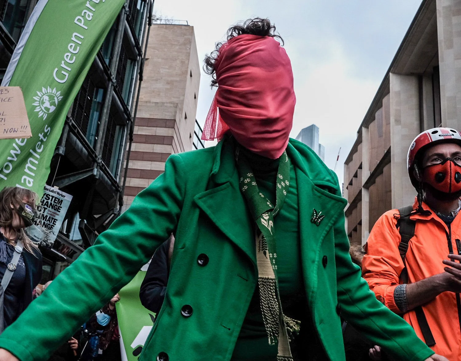 Protesters marching in a city street during a climate change demonstration, with one person in a green coat and red headscarf, and another person in an orange jacket and helmet.