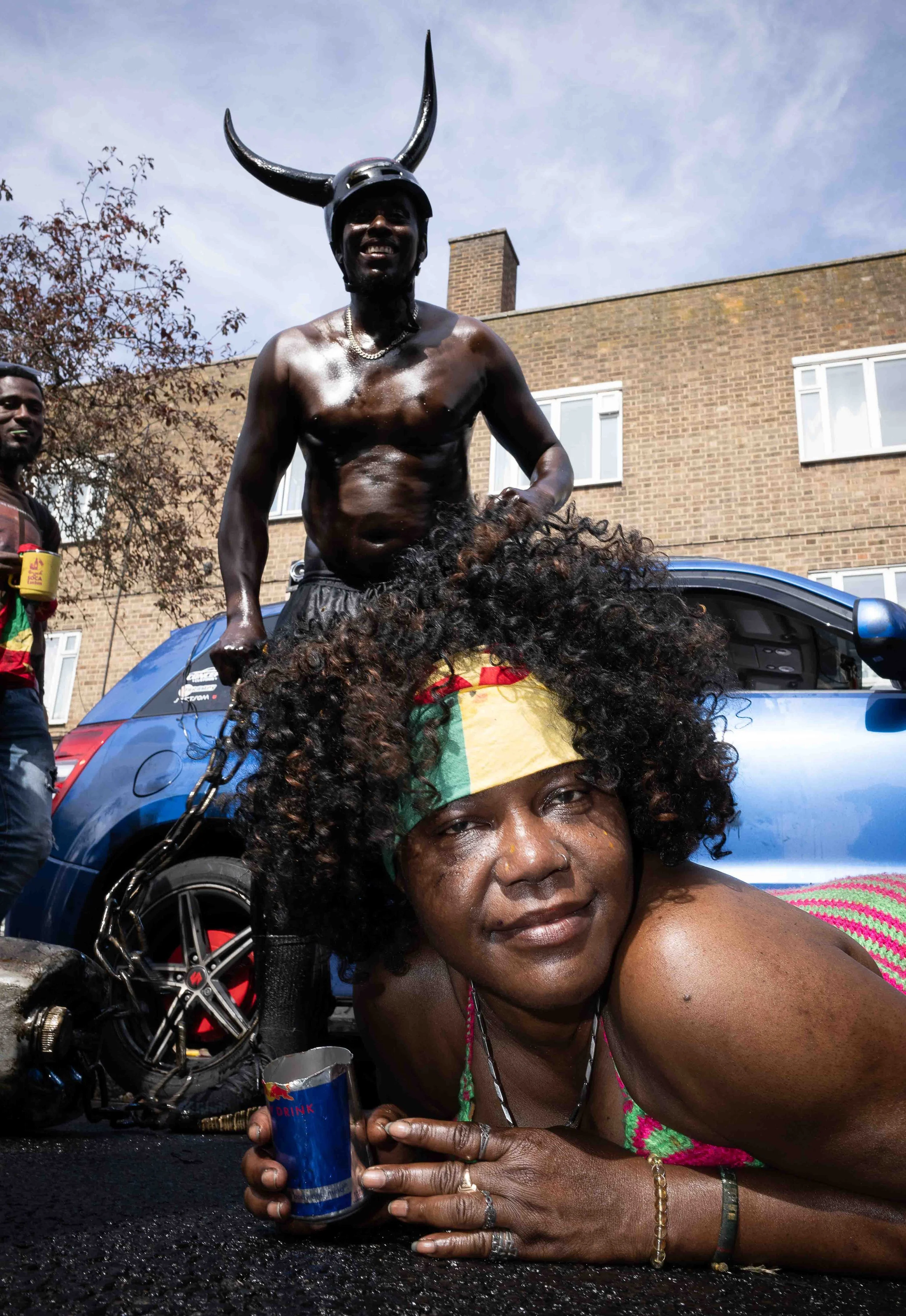 A person wearing a Viking helmet with large horns, shirtless, standing next to a person with curly hair wearing a colorful headband, lying on the ground and holding a can. A motorcycle and a house are visible in the background.