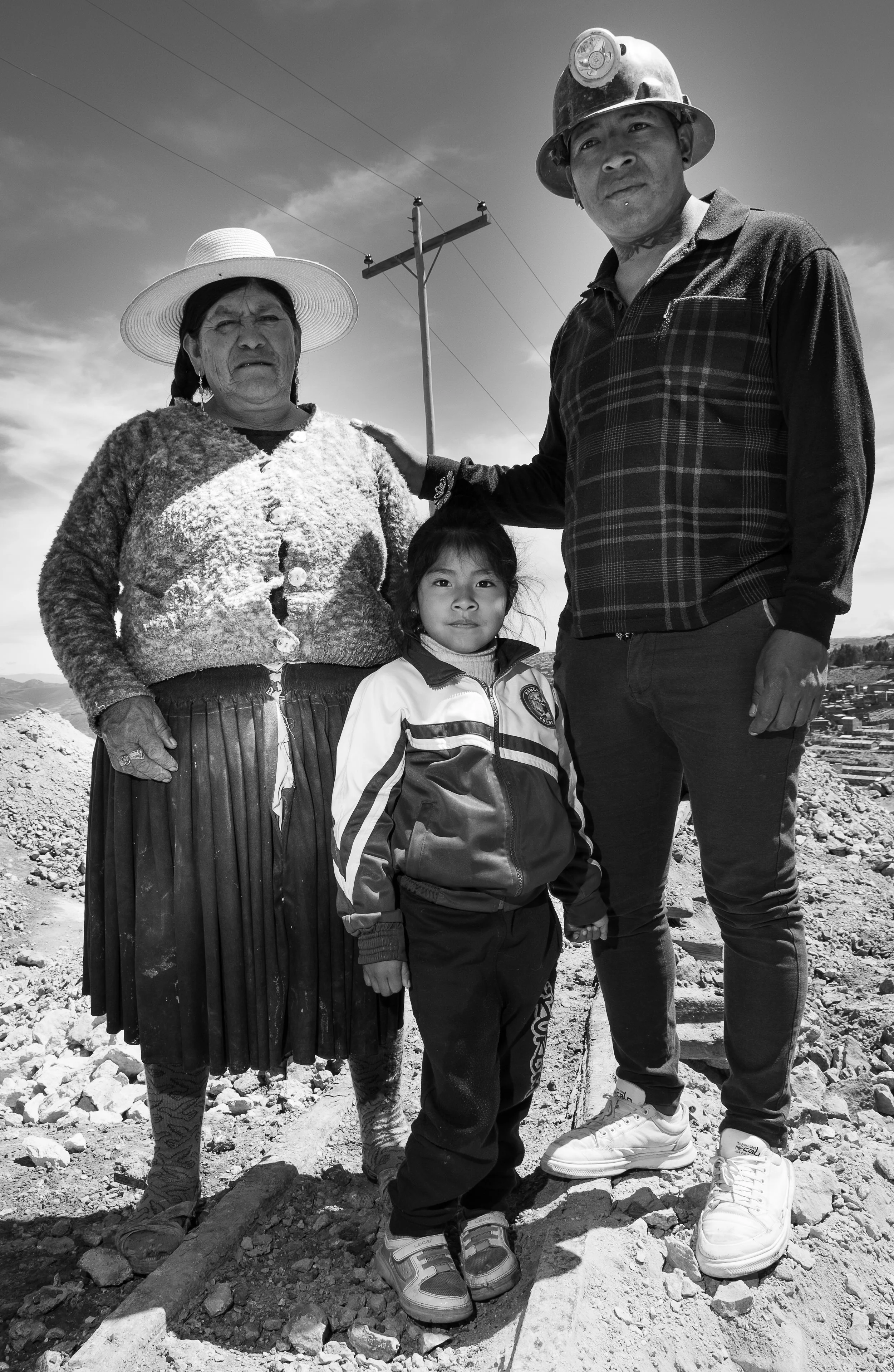 A family of four standing outdoors on a rocky terrain under a utility pole, with a cloudy sky background, in black and white.