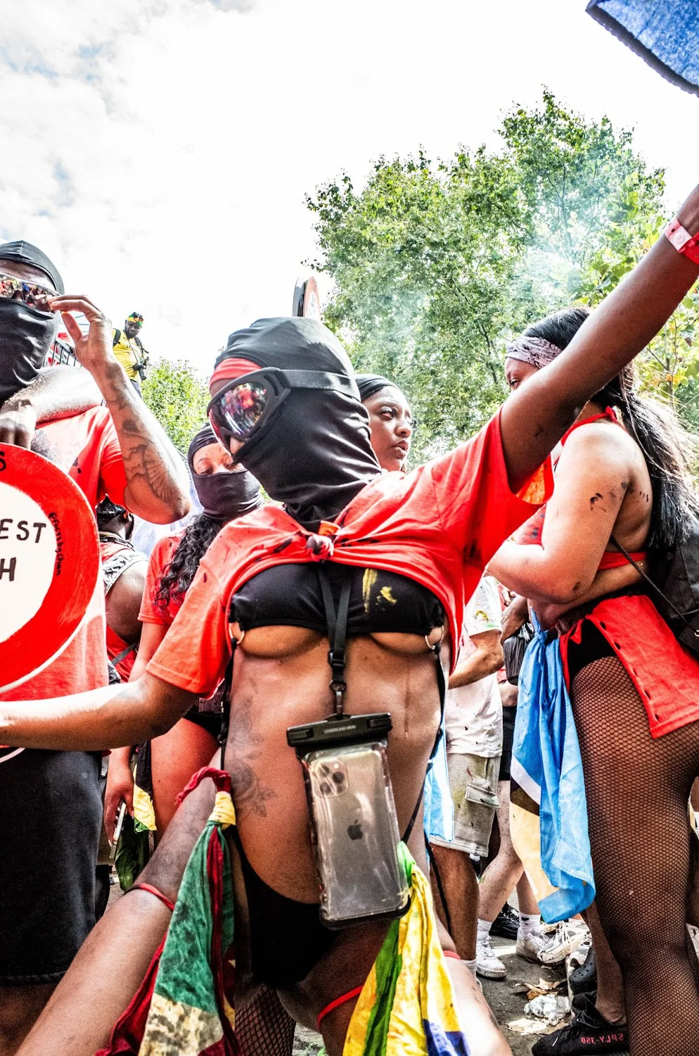 A group of protesters, many wearing red clothing, some with their faces covered, gathered outdoors during the daytime, with trees and cloudy sky in the background.